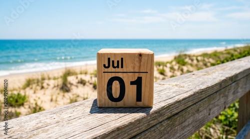 A wooden calendar block displaying July 01 on a table with a blurred beach and ocean in the background under clear sky.
