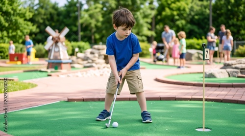 Young boy playing miniature golf on a sunny day. Focused child holding putter and aiming at golf ball on green artificial turf. Outdoor family leisure activity
