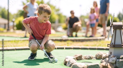 Happy young boy crouching with putter on miniature golf course. Smiling child playing mini golf while family watches in background. Outdoor summer leisure activity