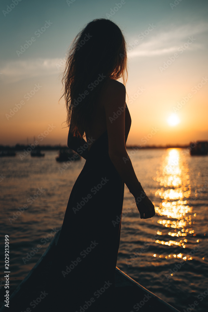 Fototapeta premium Woman silhouette standing on boat during golden hour sunset, Female traveler on water yacht at sunrise with elegant dress