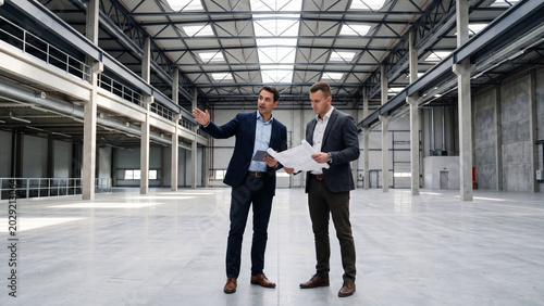 Two professional businessmen discussing construction plans inside a large empty industrial warehouse building