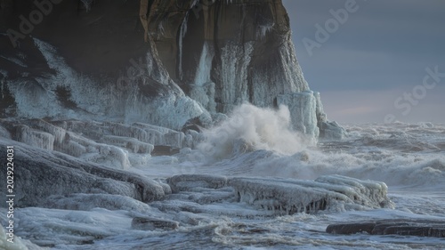 Dramatic coastal scene with icy cliffs crashing waves and snowy shoreline during stormy weather