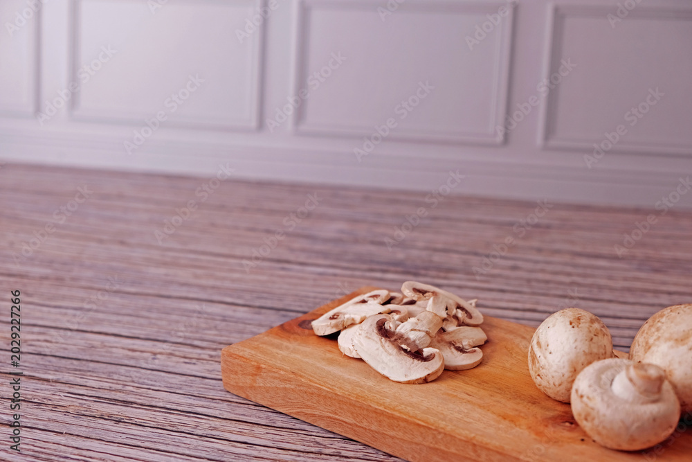 Fototapeta premium Fresh champignon mushrooms sit on a rustic wooden board. Sliced pieces rest beside whole caps, softly lit. Wooden surface shows aged grain and texture. Natural, wholesome, and simple. Ideal for health
