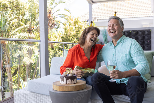 Senior couple celebrating on balcony, red blouse, blue shirt, holding wine and gift, cake with 50