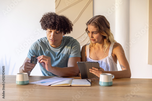 Couple sitting at wooden table in bright kitchen examining credit card, tablet and paperwork