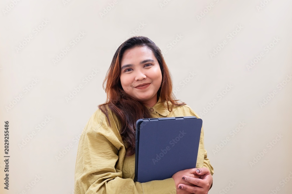 Fototapeta premium Warm smile radiates from a confident woman holding a clipboard. Her hands grip the folder, poised for work or presentation. Soft beige backdrop highlights her calm, professional demeanor.