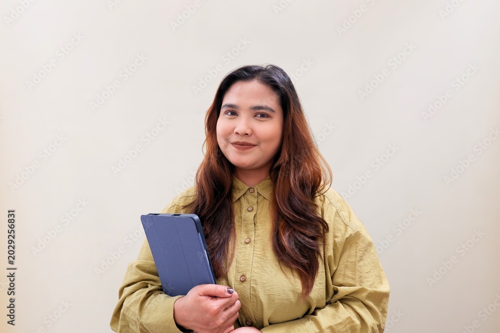 Fototapeta premium Warm smile radiates from a confident woman holding a clipboard. Her hands grip the folder, poised for work or presentation. Soft beige backdrop highlights her calm, professional demeanor.