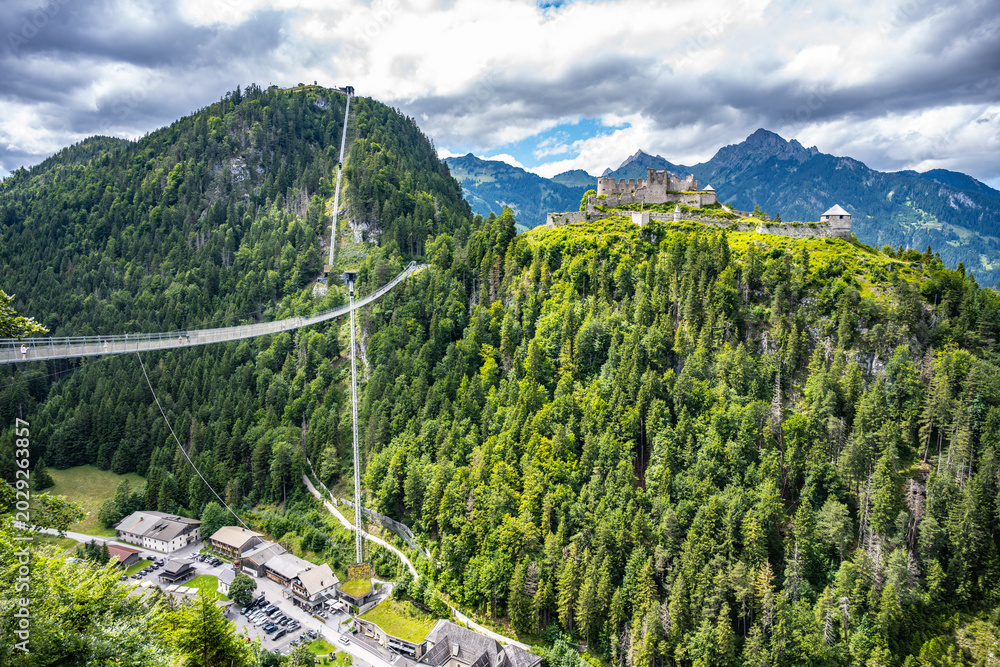 Fototapeta premium A suspension bridge connects the hillside to the ruins of Ehrenberg Castle. The Schlosskopf fortress stands above. Visitors can explore the area surrounded by mountains and trees.