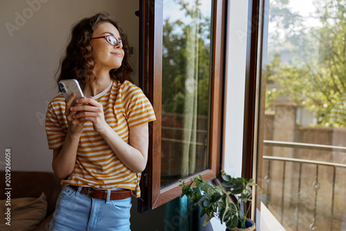 Valokuva Smiling woman with curly hair standing by an open window, wearing a striped shirt and jeans