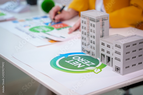 Close-up of sustainable real estate planning with building models on desk and net zero energy symbol, person drafting eco design in office.