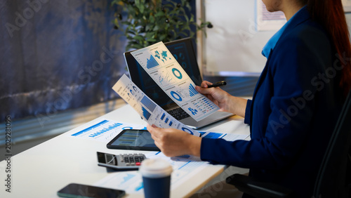 Businesswoman in office reviewing printed analytics reports with charts and graphs, using tablet and laptop on desk with calculator and coffee.