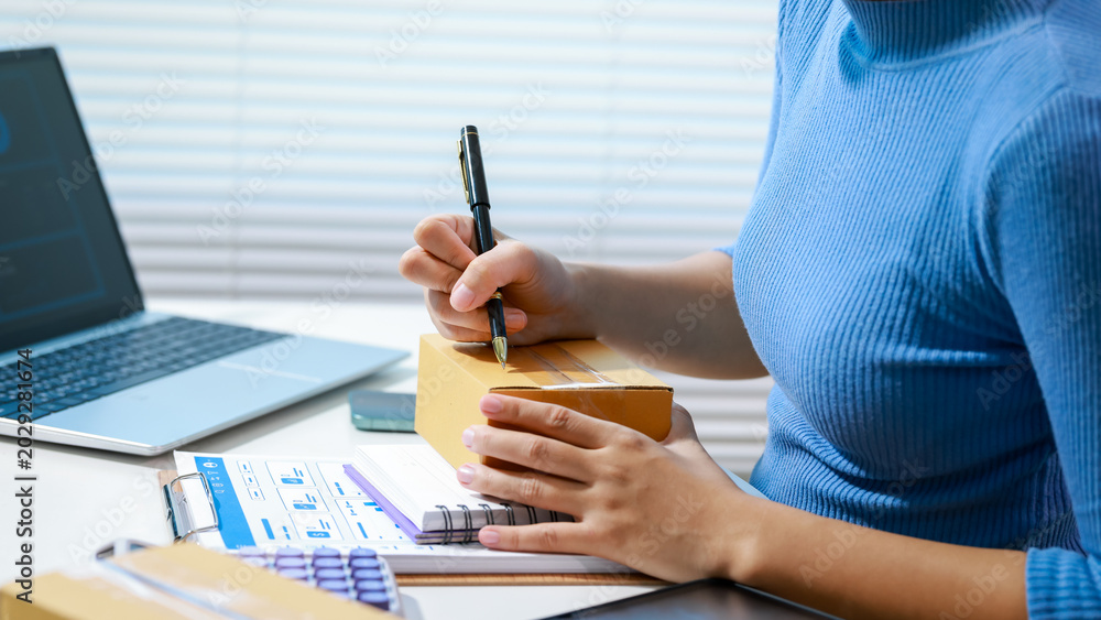 Fototapeta premium Person at desk writing on shipping box beside laptop, paperwork, calculator, and smartphone, suggesting small business order fulfillment and logistics in home office.