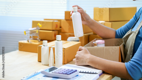 Tableau sur toile Small business owner packing skincare bottles into shipping boxes at home, with calculator and paperwork on table, e-commerce fulfillment and inventory concept
