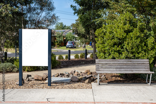 Obraz na plátně Blank white mockup sign on metal pillars next to a public park bench on a pavement, with a residential suburban street, trees in the background