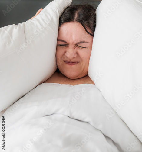 Fotografie Middle-aged Caucasian woman with brown hair is lying in bed, covering her ears with white pillows