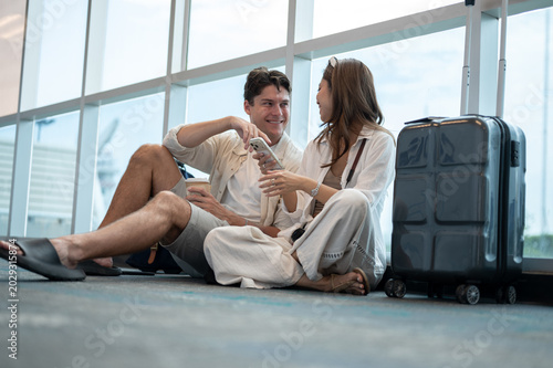 Asian woman girlfriend talking to white man boyfriend while sitting with luggage on floor in airport