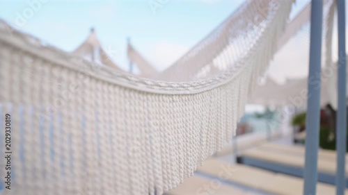 Close-up of elegant white cotton hammock with decorative fringe swaying gently at a beach resort in Santorini Greece. Soft focus background reveals thatched umbrellas and blue sky on a sunny day.
