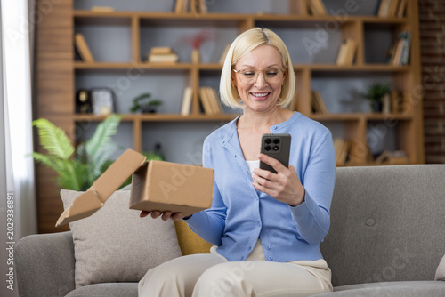 Canvastavla Senior woman sitting on a sofa, smiling and holding a delivery box while reviewi