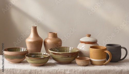 A collection of handcrafted ceramic bowls, mugs, and vases in earthy tones displayed on a table with natural light and shadows.