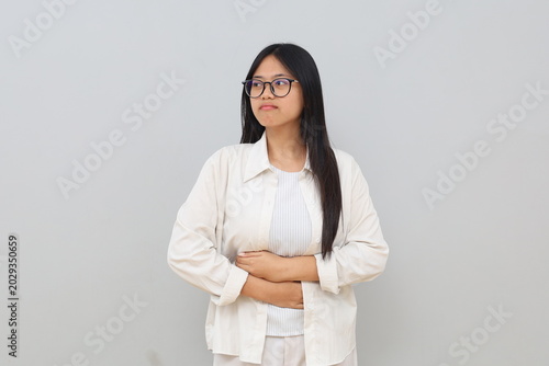 A young Asian woman is having stomach ache, bending over and holding her stomach, feeling uncomfortable due to menstrual cramps, stomach ache, diarrhea, standing isolated on white background.