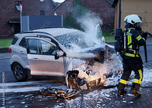 Firefighters extinguishing burning passenger car with foam during emergency response operation on residential street