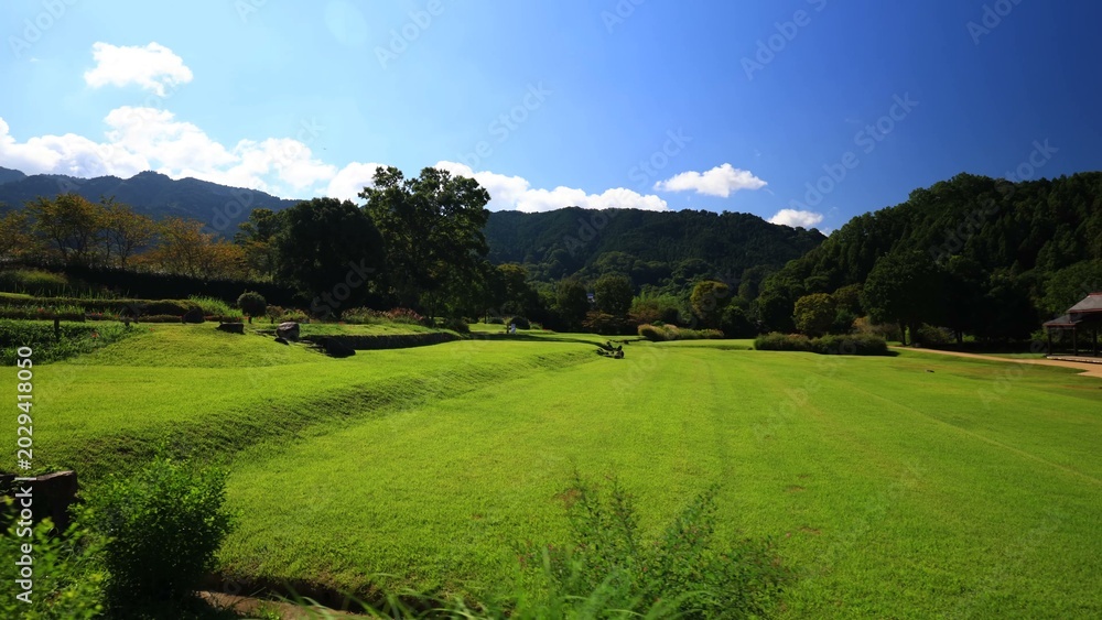 Obraz premium A spacious grassy field stretches toward forested hills with two open wooden pavilions standing in sunlight beside a winding dirt path creating a calm natural setting of balance and rural simplicity 