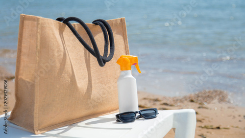 Stylish eco beach bag with sunscreen spray sunglasses near sparkling sea shoreline, capturing authentic vacation mood, summer travel lifestyle, relaxation, sustainable tourism concept copyspace