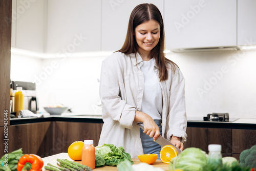 Young woman standing in modern kitchen, smiling while cutting fresh oranges and preparing a healthy, nutritious meal