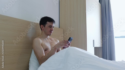 Young shirtless caucasian man sitting in bed under white sheets, texting on his mobile phone and browsing social media after waking up in his bright bedroom or a modern hotel room