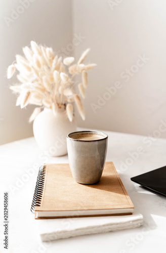 Coffee cup on a notebook beside a laptop, minimalist home office still life with soft light