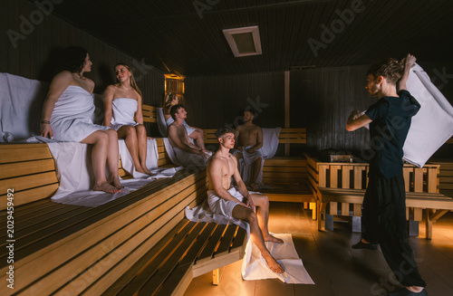 Male sauna master waving a towel during an aufguss ritual for a group of young people in a large dry sauna