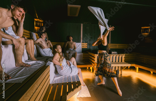 Female sauna master waving a towel during an aufguss ritual for a group of young people in a dry wooden sauna