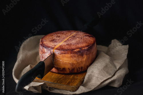 A cheesemaker cuts a wheel of hard cheese with a knife. Exquisite cheese in wine on a wooden board. A farm product made from milk on a dark background. Delicious hard cheese. Wheel of cheese. Perfect 