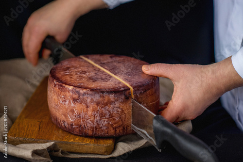 A cheesemaker cuts a wheel of hard cheese with a knife. Exquisite cheese in wine on a wooden board. A farm product made from milk on a dark background. Delicious hard cheese. Wheel of cheese. Perfect 