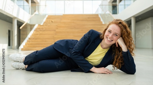 Businesswoman with red hair and a dark blue suit lying on the floor in a bright corporate office, smiling broadly while showcasing genuine happiness and relief from work pressure
