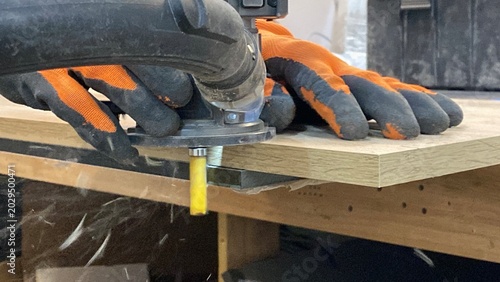 Fotografie Close-up of a worker in protective gloves using a hand router to trim the edge of a wooden board in a workshop