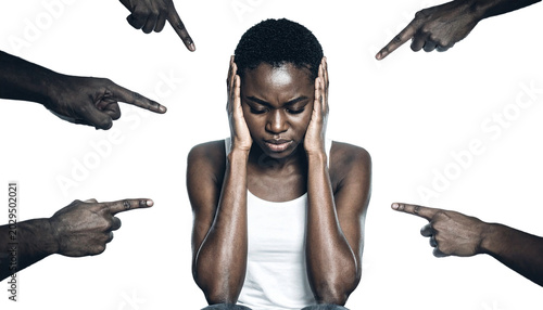 A person sits, head down, hands over ears, surrounded by accusing fingers pointing