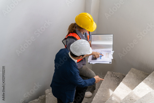 Professional engineers wearing hardhats holding document folder to discuss and inspect staircase construction progress inside residential building site from high angle view