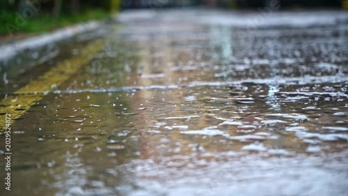 Close up of raindrops hitting wet concrete pavement in city street. Outdoor urban scene with shallow focus, puddles, ripples, and reflections during rainy weather.