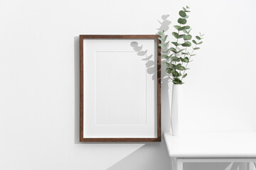 Blank vertical photo frame with dark wood border on white wall next to a tall white vase containing fresh eucalyptus stems in a minimalist interior