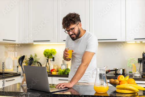 Arabian man drinks orange juice in home kitchen while reading news on laptop for morning start. Young guy follows healthy vitamin breakfast routine and boosts energy with citrus hydration before work.