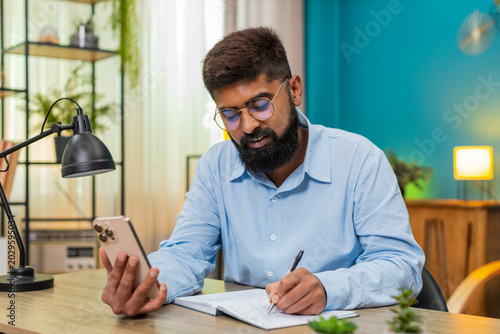 Indian man at home attends smartphone lecture nods and writes notes in notebook discipline focused. Arabian guy at desk stays focused tapping screen following teacher maintaining solid remote study.