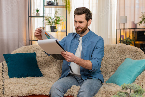 Φωτογραφία Man at home sits on the sofa, anxiously flipping through bills and documents searching for something crucial