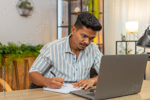 Canvas Print Indian man at home studies online course with laptop and notebook writes notes completes test