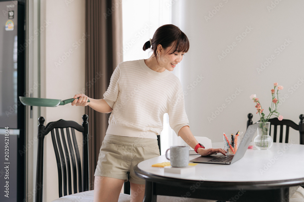 Obraz premium One cheerful young asian woman enjoys multitasking in beautiful home kitchen holding cooking pan and typing on modern laptop while smiling and searching for online recipe