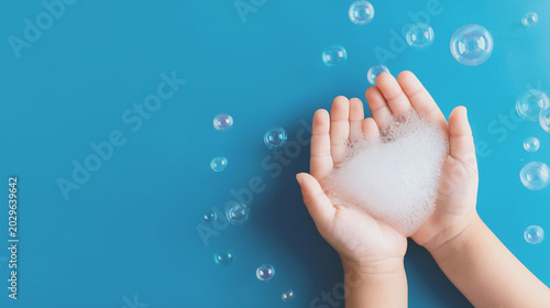 Cute little baby's hands covered with soft soap bubbles resting on a pastel blue background.