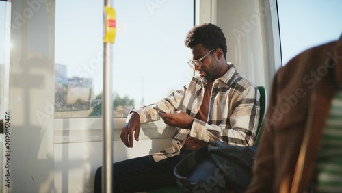 African American man with beard and glasses sitting by window on bus using smartphone. Passenger leaning back relaxed scrolling screen during sunny day ride. Casual moment on public transport.