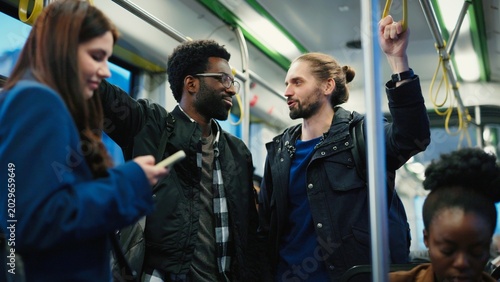 Fotografie Man with glasses and bearded friend smiling while standing inside tram talking to female colleague in blue jacket