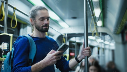 Caucasian man with beard wearing blue sweater standing on city bus holding pole with one hand and smartphone in other. Male reading online news during evening commute home from work.