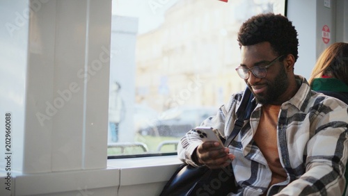 Smiling African American man with beard wearing glasses holding smartphone while sitting by bus window. Passenger checking messages during ride looking pleased. Relaxed moment on public transport.